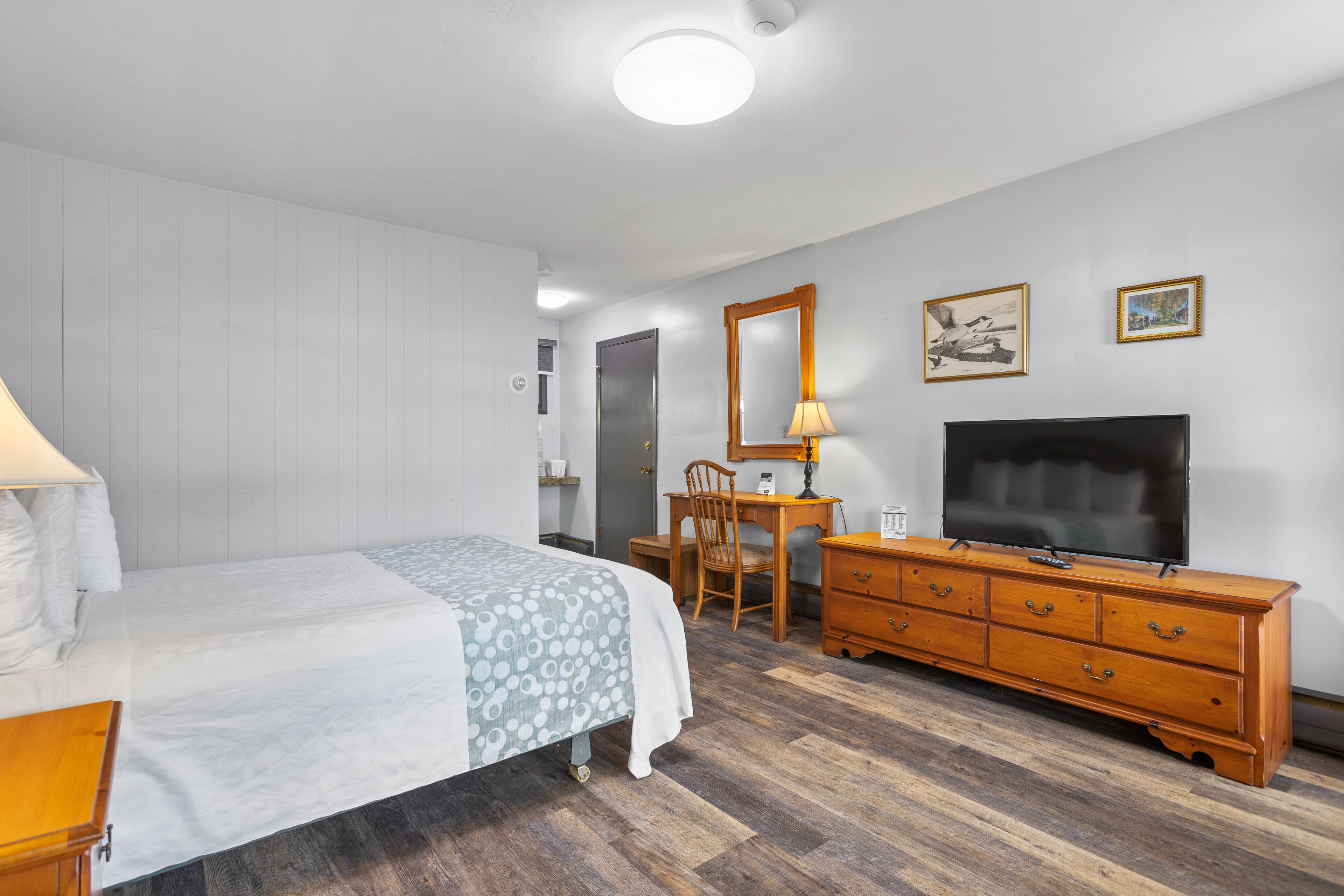 Un dormitorio con cama, cómoda de madera, escritorio con silla, televisor y espejo.