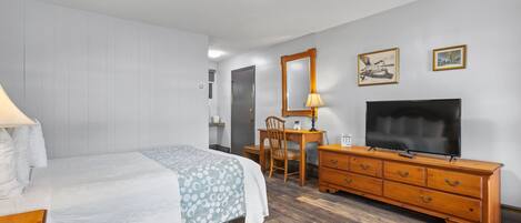 Un dormitorio con cama, cómoda de madera, escritorio con silla, televisor y espejo.