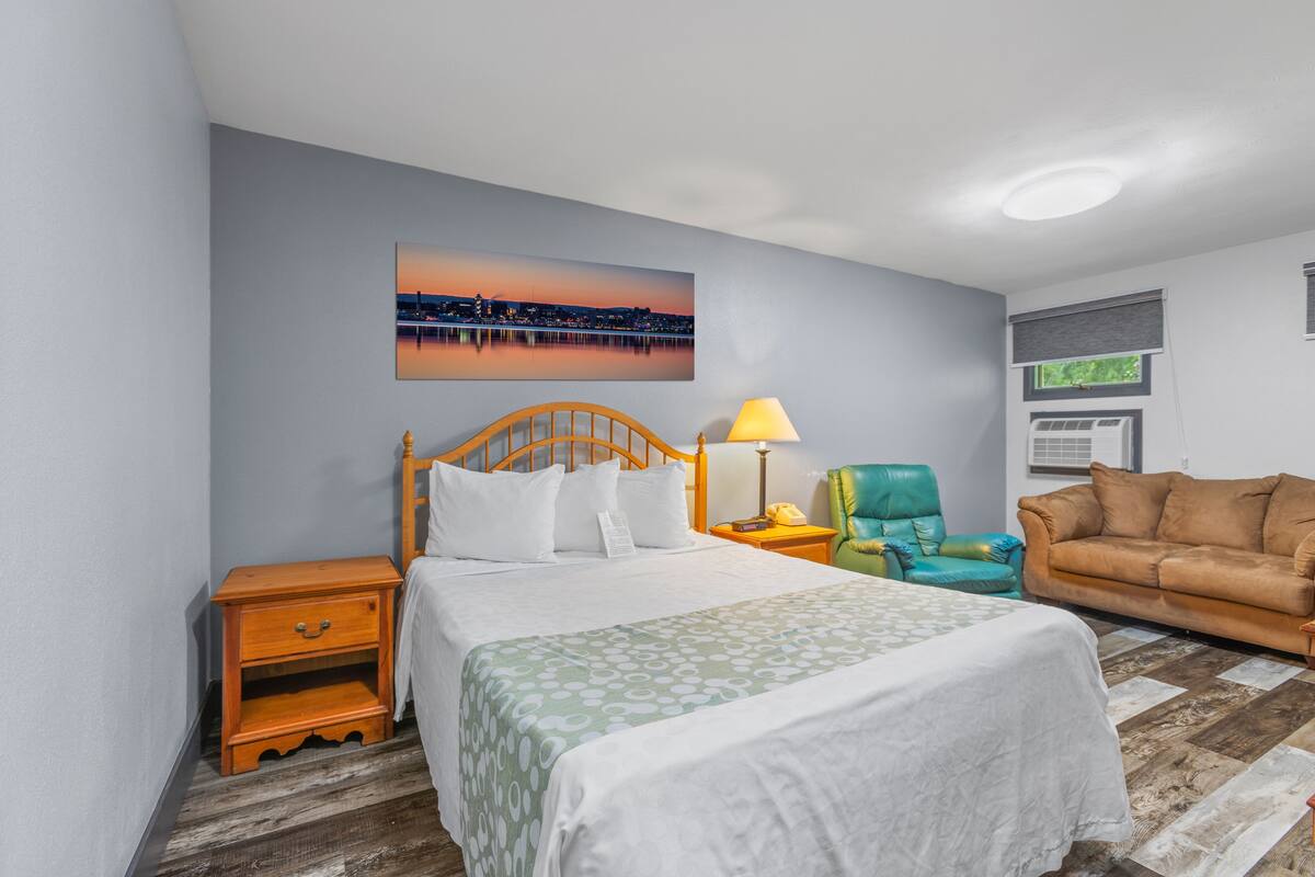 A hotel room with a bed, a nightstand, a green armchair, a brown sofa, a cityscape painting, and a window with blinds.