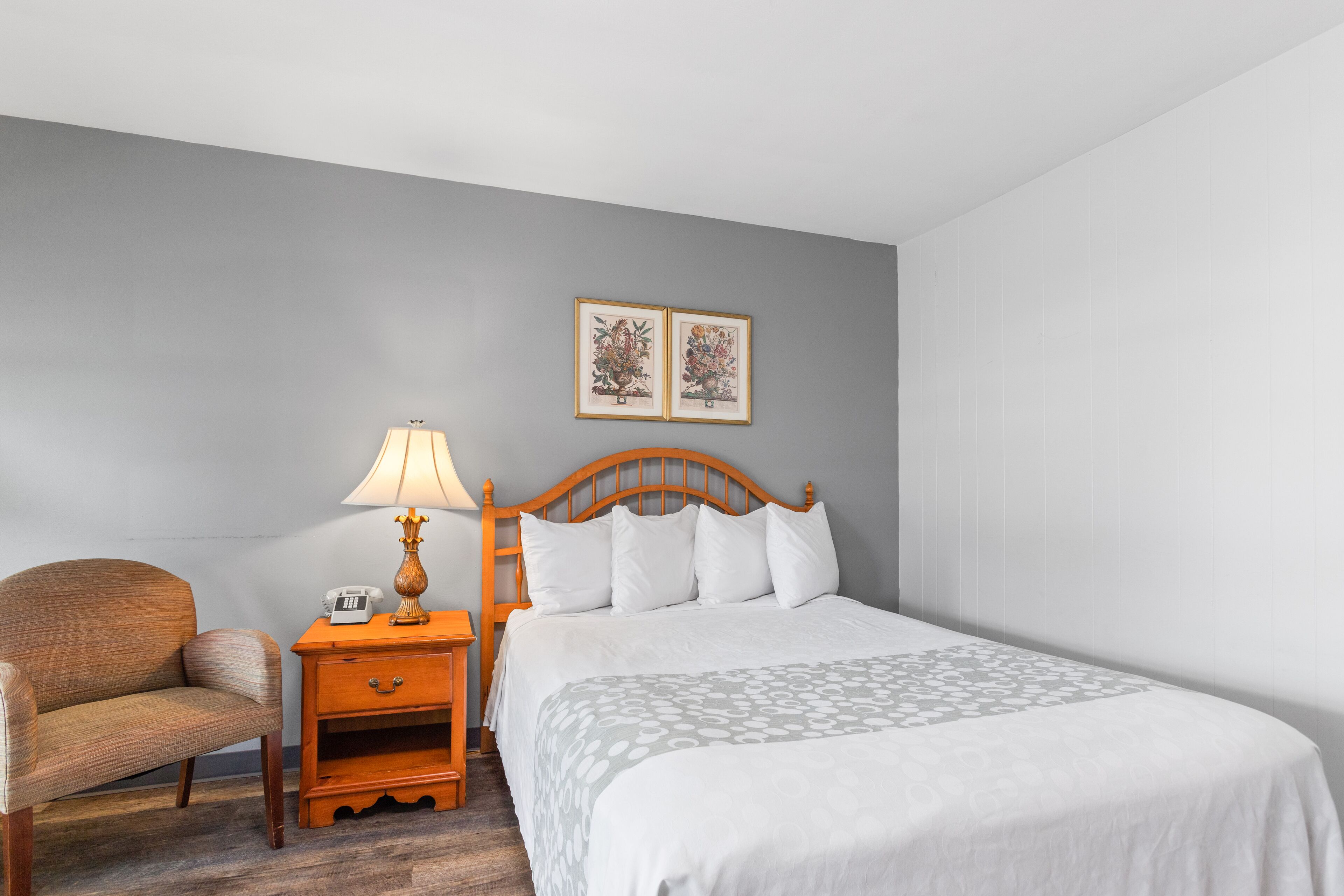 A neatly arranged hotel room with a bed, a chair, a nightstand with a lamp, and two framed pictures on the wall.