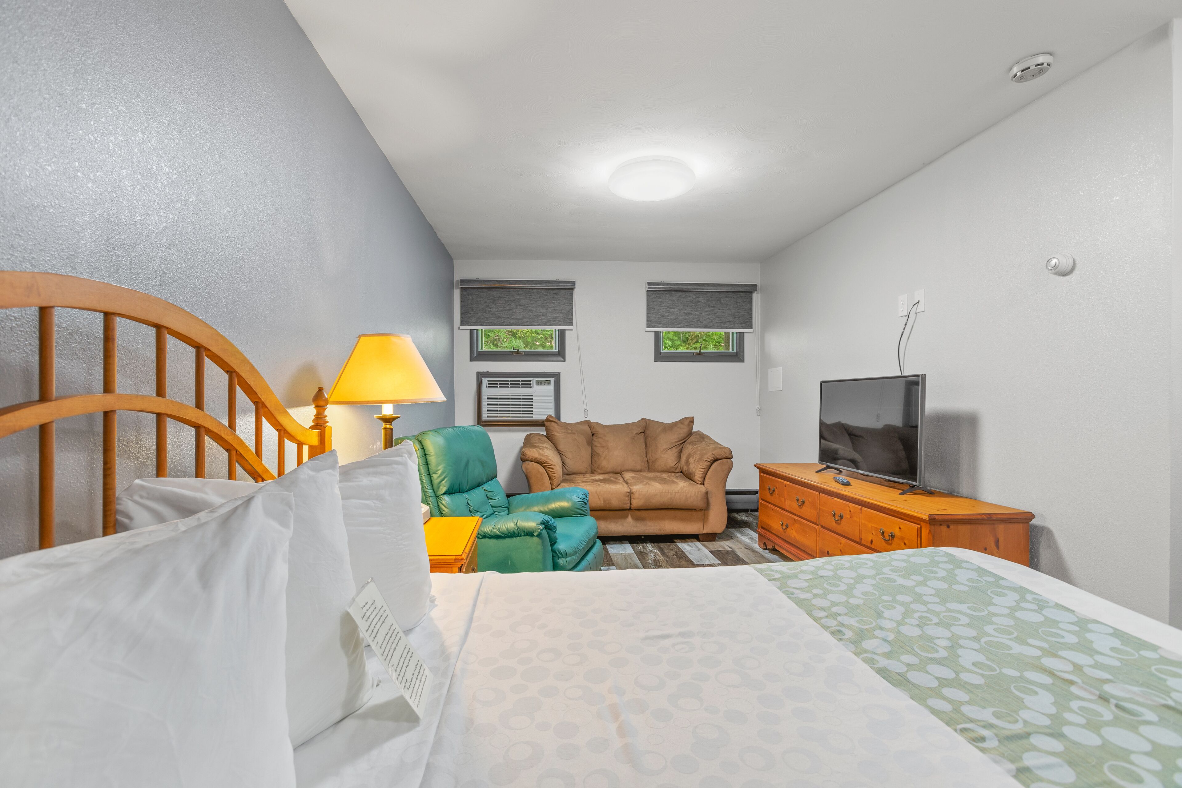 Una habitación de hotel con una cama, un cabecero de madera, un sillón verde, un sofá marrón, un aparador de madera con un televisor y dos ventanas con persianas.