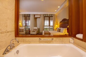 A hotel bathroom with a large bathtub, a mirror, and a view of the balcony and room.