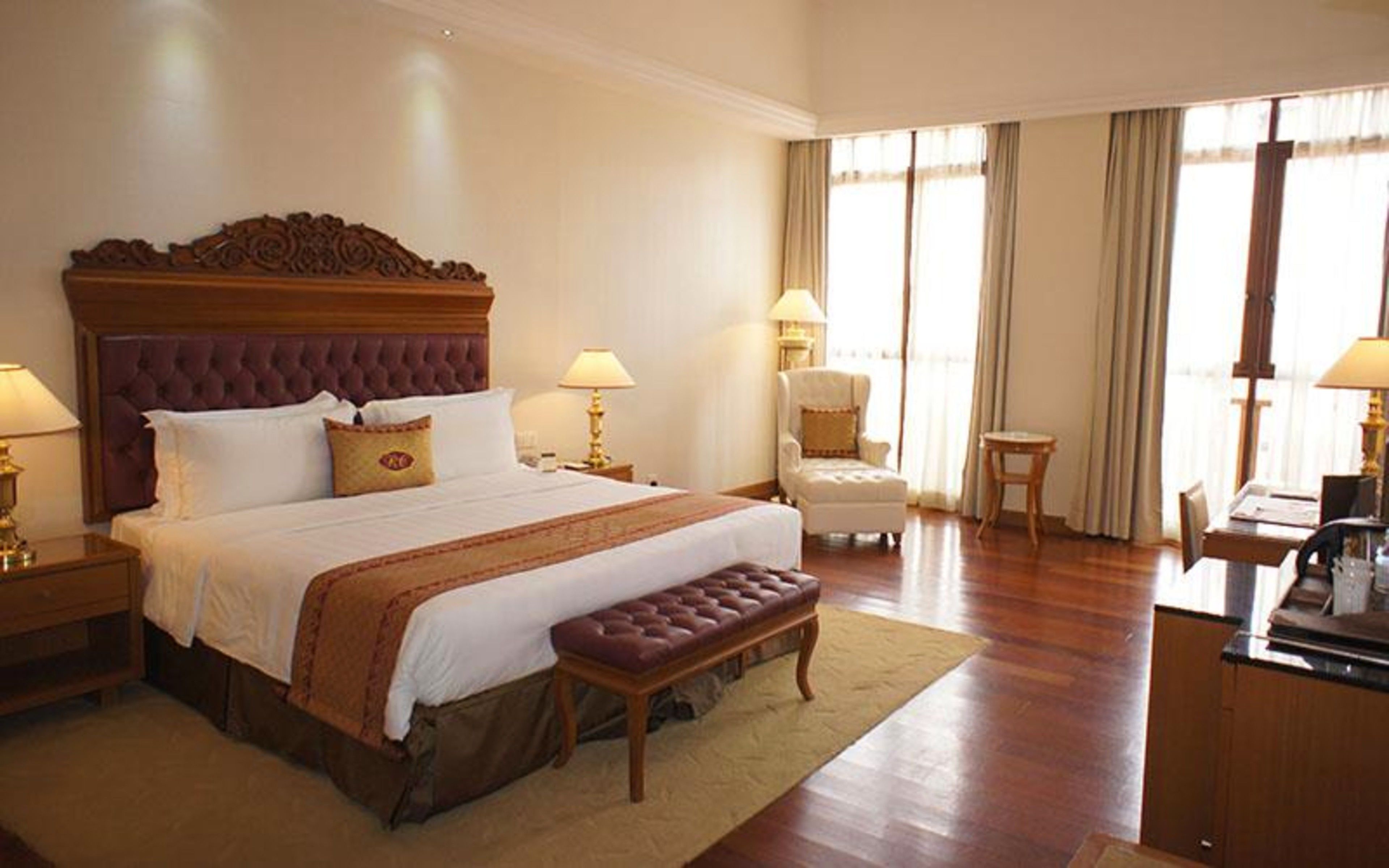 A hotel room with a large bed, a wooden headboard, two bedside tables with lamps, a chair, a small table, and a desk with a computer.