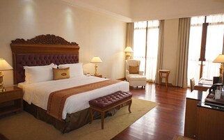 A hotel room with a large bed, a wooden headboard, two bedside tables with lamps, a chair, a small table, and a desk with a computer.