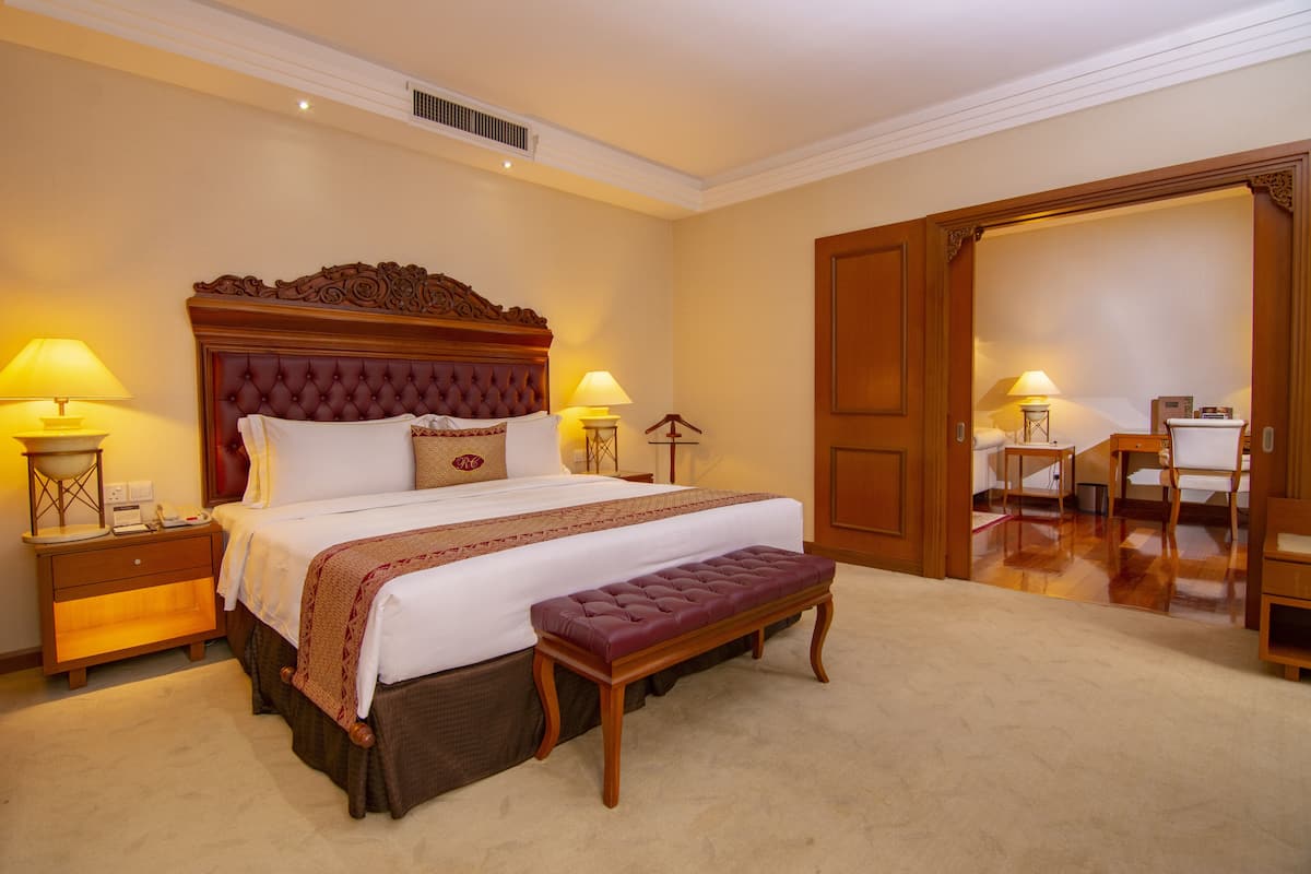 A hotel room with a large bed, two bedside tables with lamps, a wooden bench, and a wooden door leading to another room.