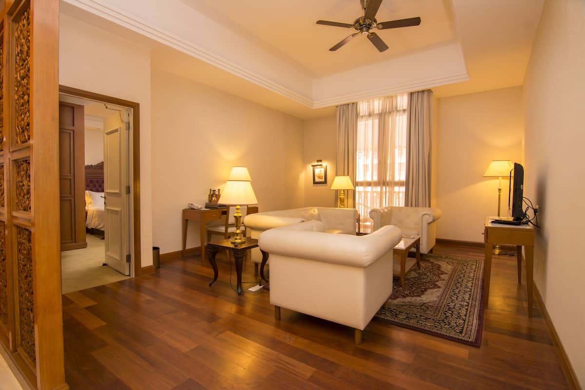 A hotel room with a wooden floor, a ceiling fan, two white sofas, a small table with lamps, and a large window with curtains.