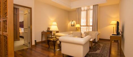 A hotel room with a wooden floor, a ceiling fan, two white sofas, a small table with lamps, and a large window with curtains.
