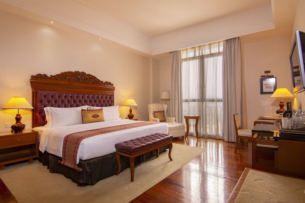 A hotel room with a large bed, two bedside tables with lamps, a wooden bench, a desk with a chair, and a large window with curtains.
