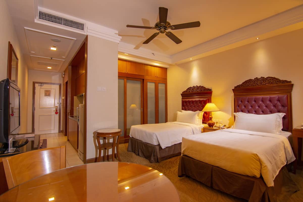 A hotel room with two beds, a dining table, a ceiling fan, and a kitchenette.