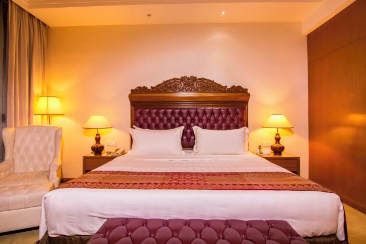 A hotel room with a large bed, two bedside lamps, a chair, and a wooden headboard.