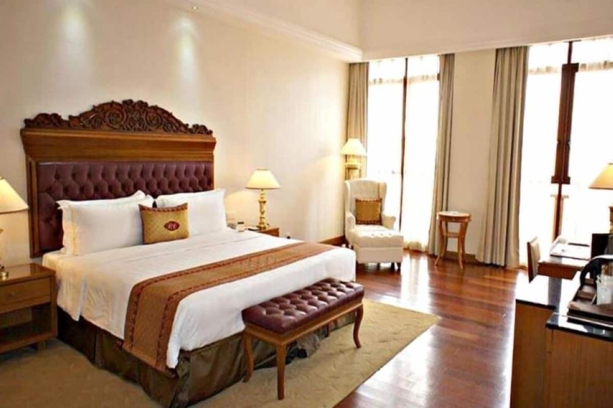 Quarto de hotel com uma cama grande, um banco, duas mesas de cabeceira, uma cadeira e uma escrivaninha com uma luminária.