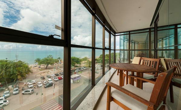A balcony with a wooden table and chairs, overlooking a beach with parked cars and palm trees. - Best Western Premier Maceió (Maceió)