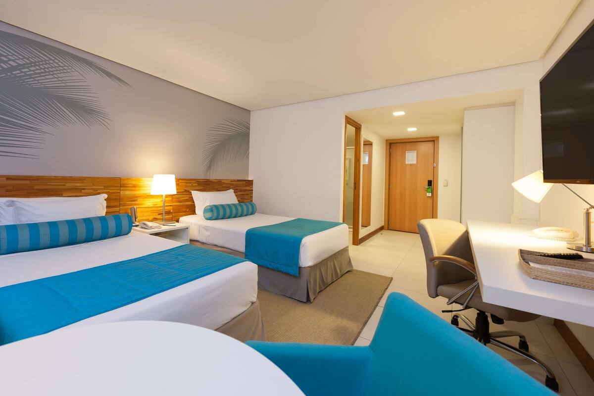 A hotel room with two beds, a desk, a chair, a TV, and a door leading to another room.
