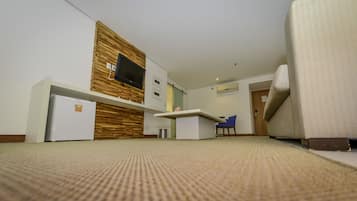 A modern hotel room with a flat-screen TV mounted on a wooden wall, a small kitchenette, a blue chair, and a sofa.