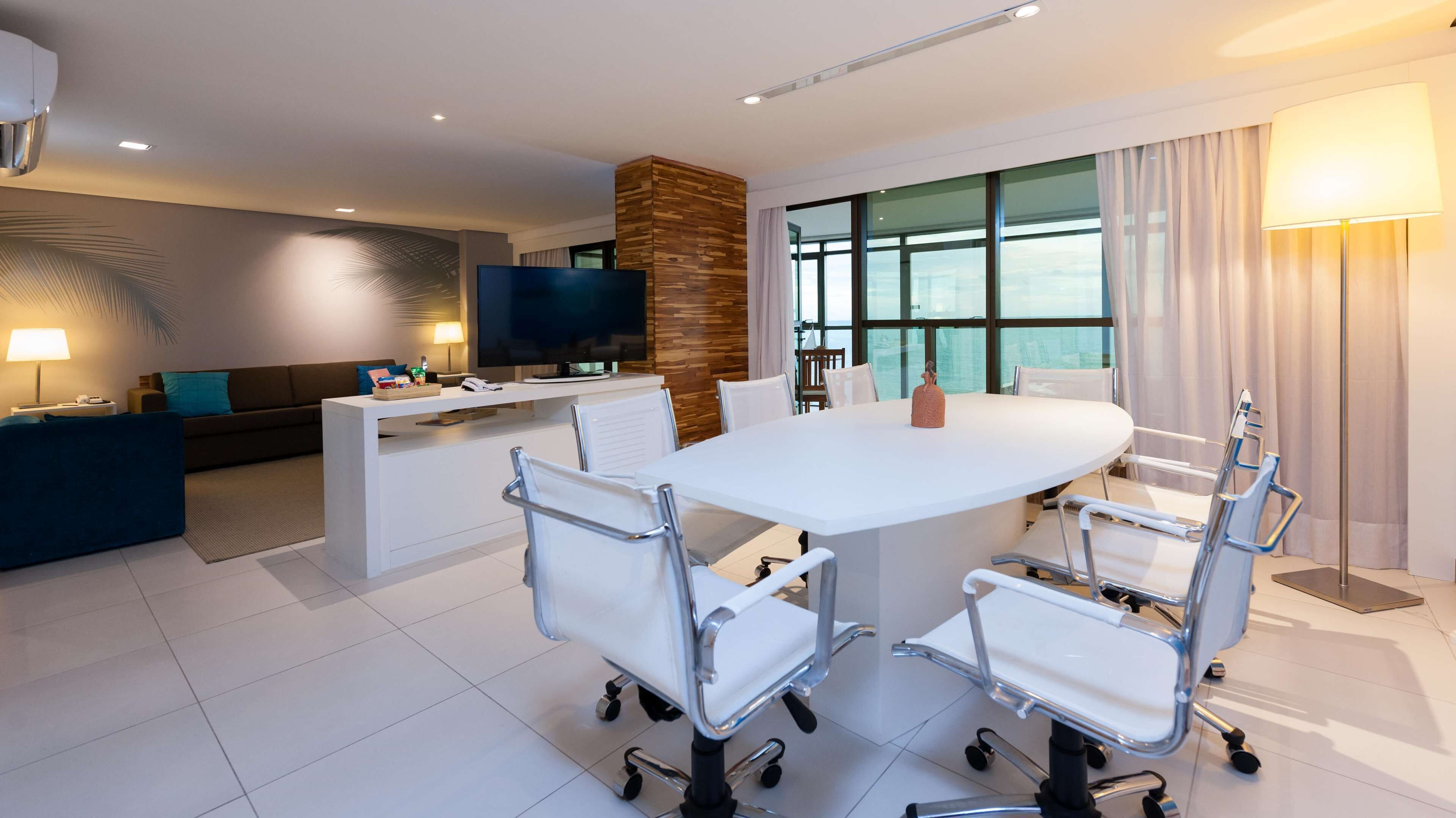 Una habitación de hotel moderna con una mesa redonda grande, sillas blancas, un televisor de pantalla plana, una pared de acento de madera y ventanas amplias.