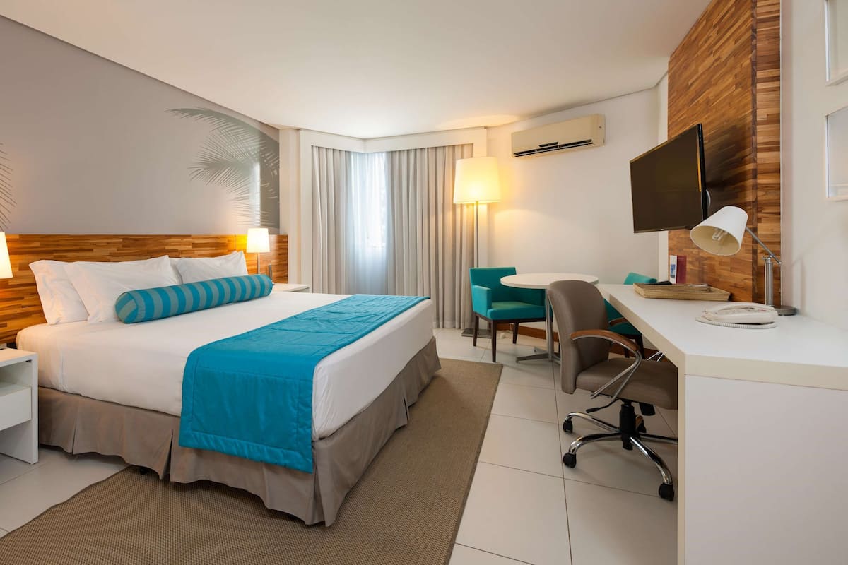 A hotel room with a bed, bedside tables, a desk with a chair, a TV, and an air conditioning unit.