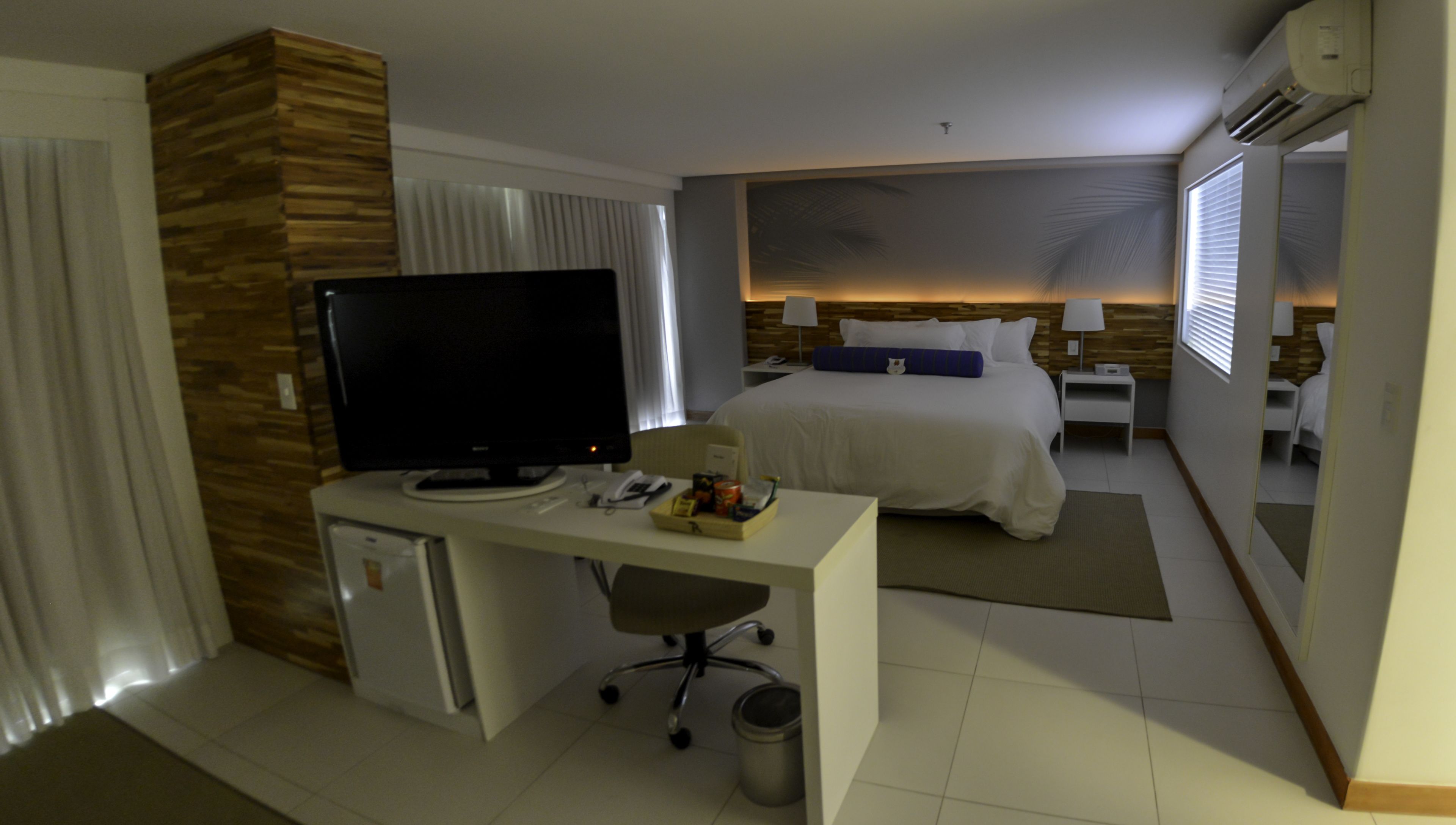 A hotel room with a bed, a desk with a television, a small refrigerator, and a bedside table with a lamp.
