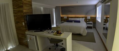 A hotel room with a bed, a desk with a television, a small refrigerator, and a bedside table with a lamp.