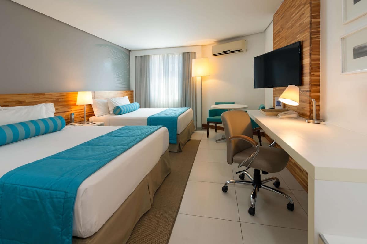 A hotel room with two beds, a desk, a chair, a TV, and a window with curtains.