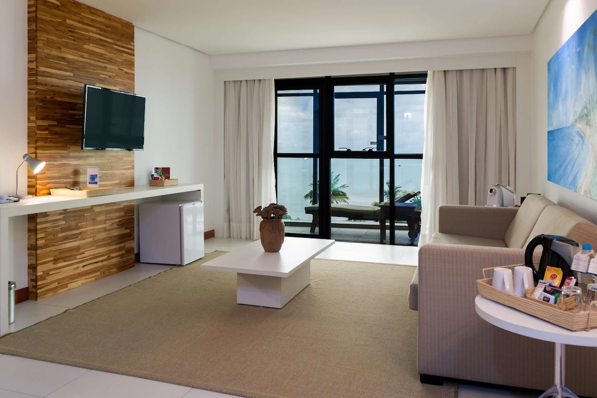 A modern living room with a flat-screen TV, a coffee table, a sofa, and a dining area with a small refrigerator.