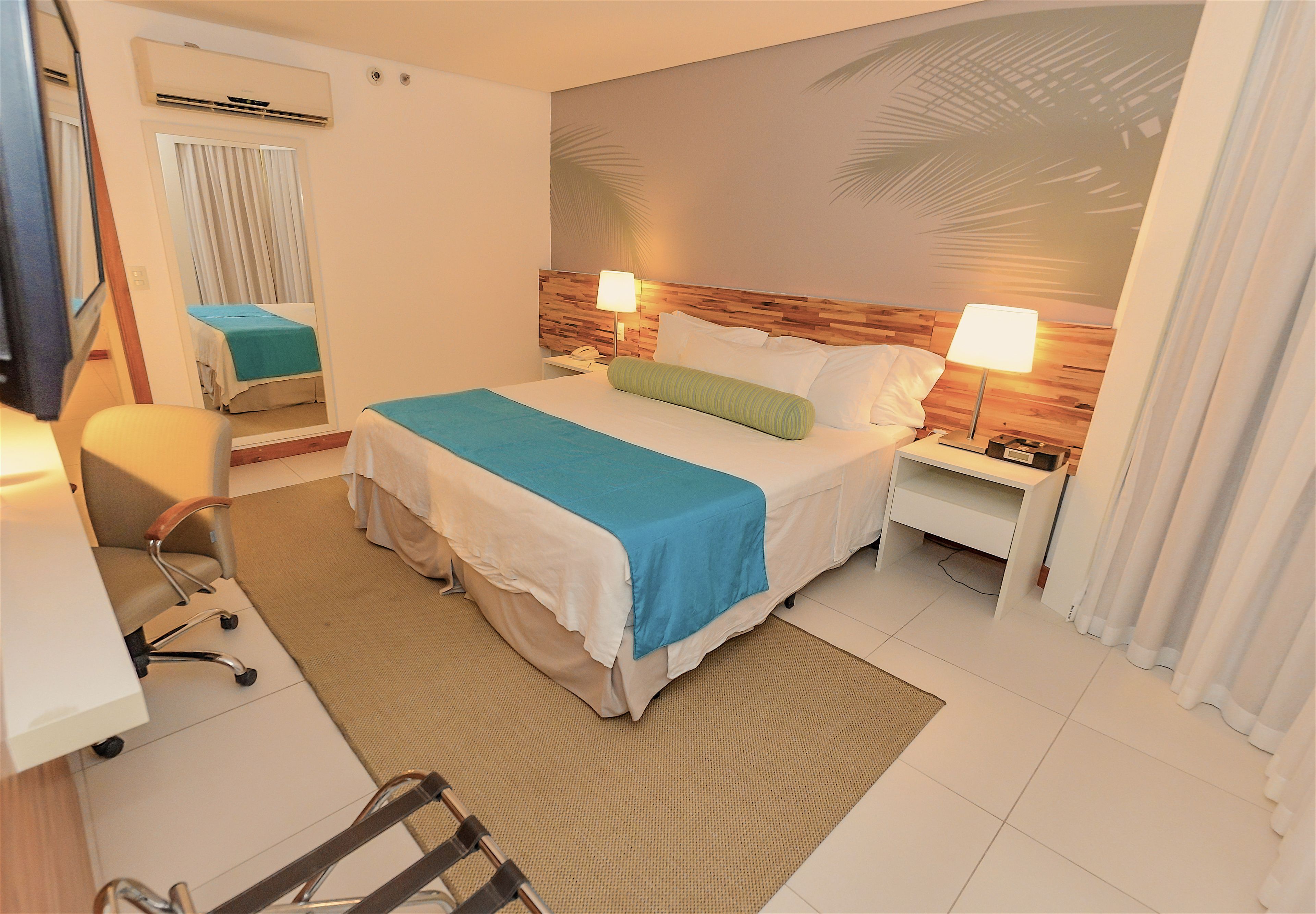 A hotel room with a bed, bedside tables, a desk, a chair, and a large mirror.