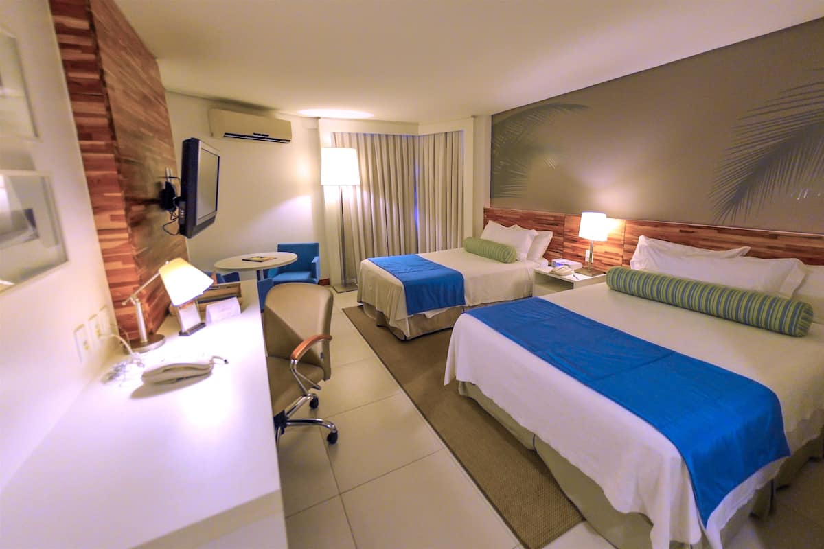 A hotel room with two beds, a desk, a chair, a small table, and a TV.