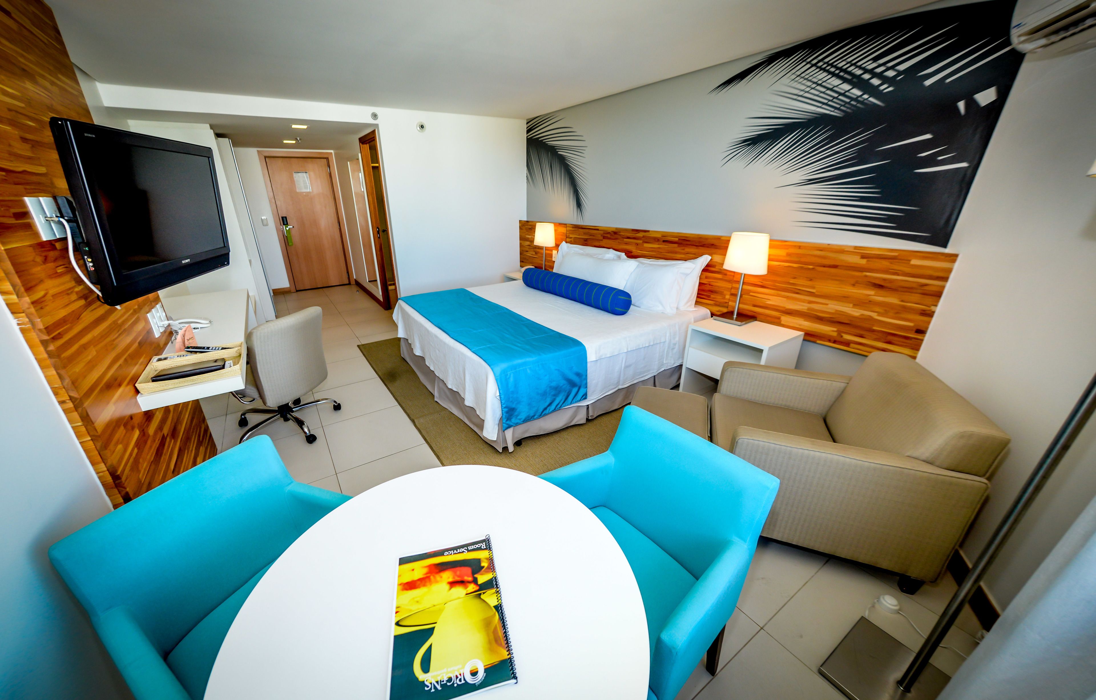 Habitación de hotel con cama, escritorio con computadora, televisor, mesa de comedor con sillas y pared con diseño de hojas de palma.
