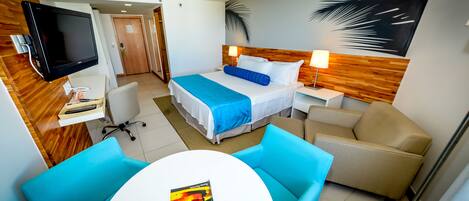 Habitación de hotel con cama, escritorio con computadora, televisor, mesa de comedor con sillas y pared con diseño de hojas de palma.