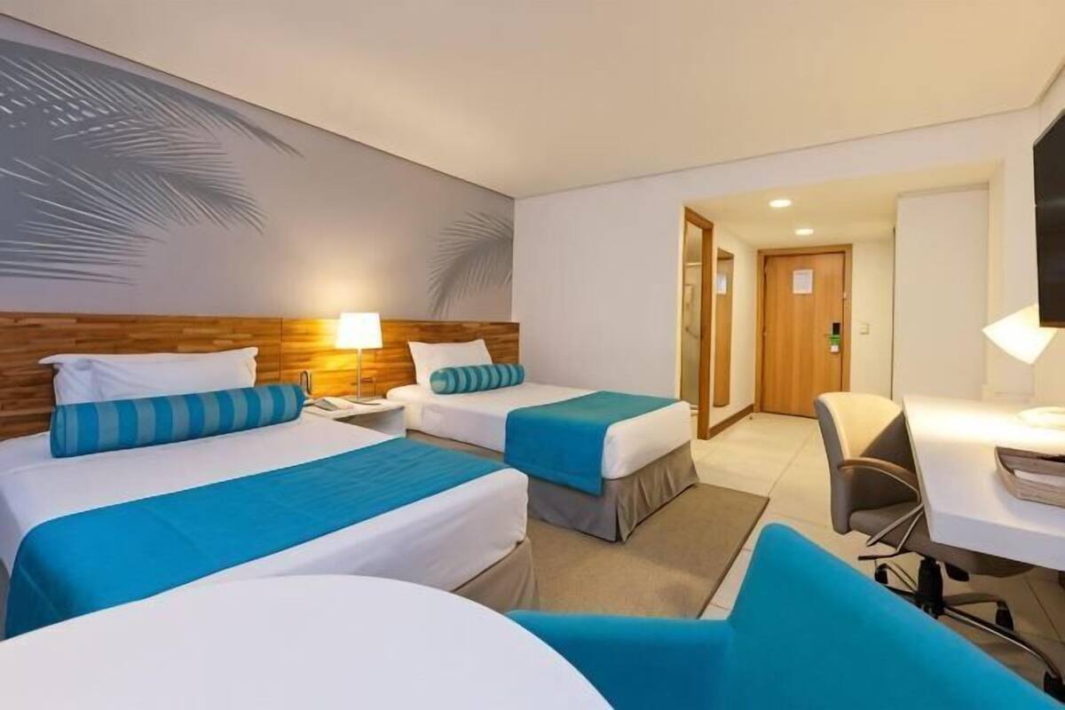 Ett hotellrum med två sängar, ett skrivbord, en stol, en tv och en dörr som leder till ett annat rum.