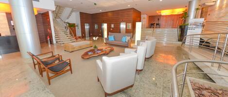 A spacious lobby with a modern design, featuring a reception desk, comfortable seating areas, and a staircase leading to an upper level.