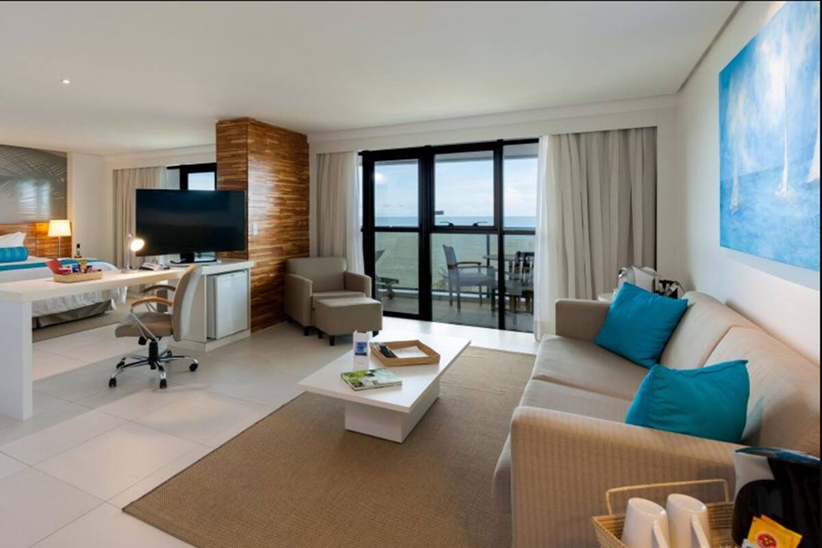 A modern hotel room with a bed, a desk, a TV, a sofa, and a balcony with a view.