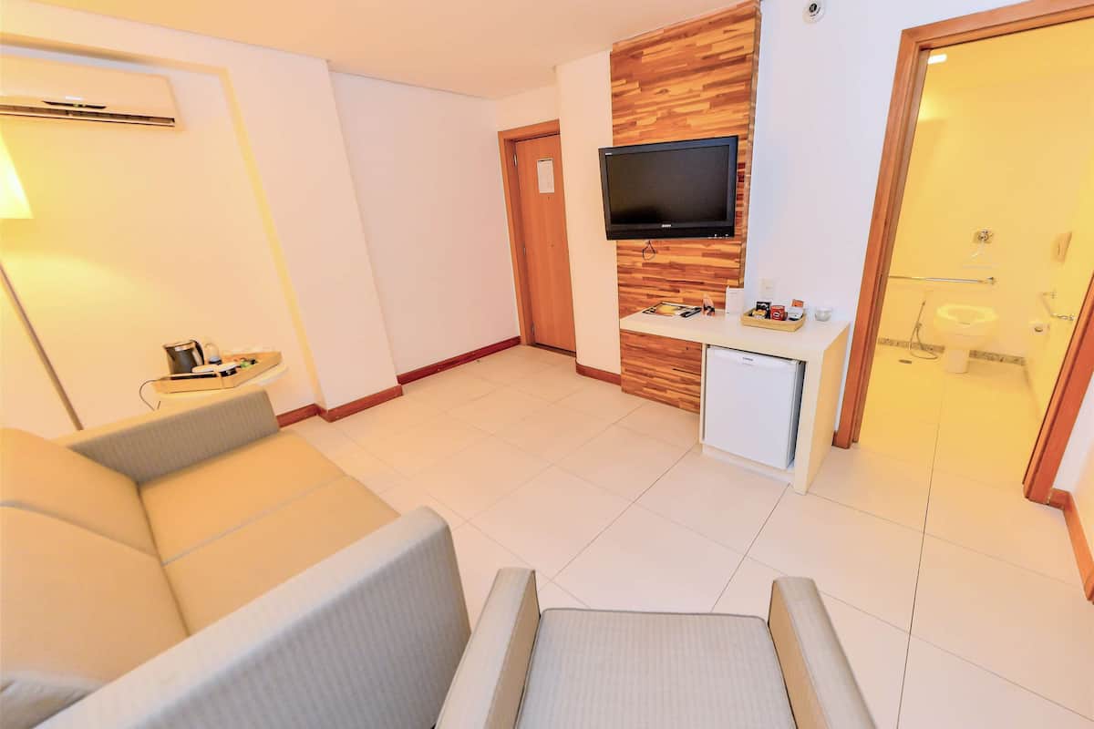 A modern hotel room with a sofa, a small desk with a TV, a mini-fridge, and a shower area.