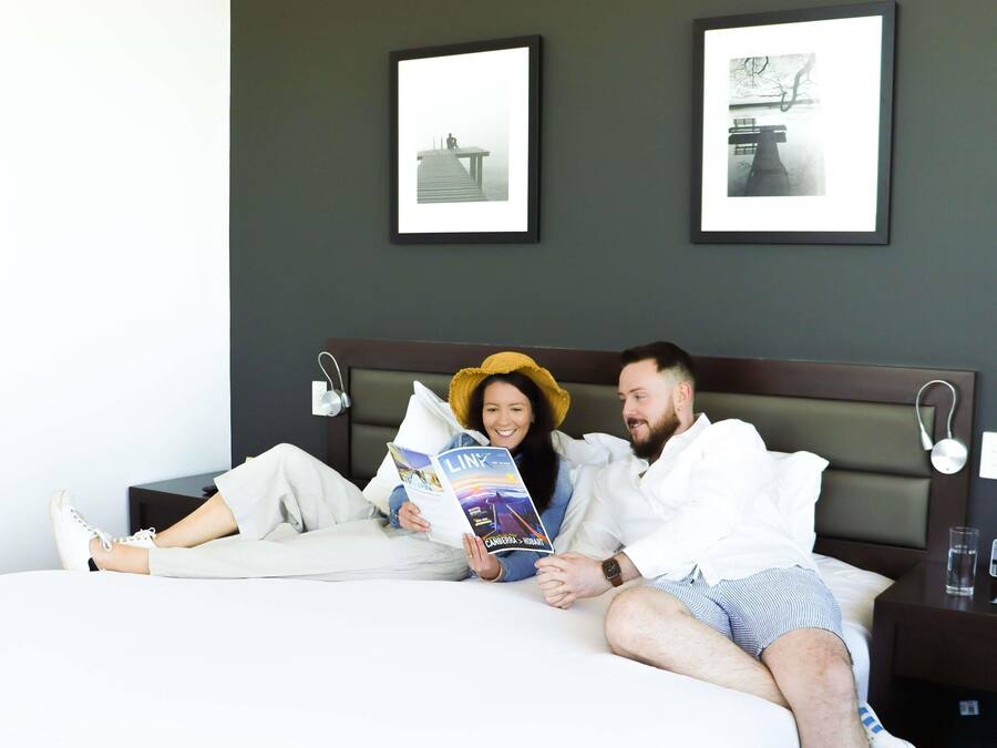 Ein Paar liegt in einem Bett mit dunkelbrauner Kopfteil und liest eine Zeitschrift, im Raum sind Bilder an der Wand.