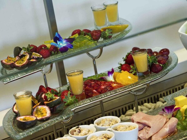 A multi-tiered glass serving tray with various fruits, juices, and a selection of meats.