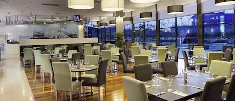 Un restaurante con varias mesas y sillas, ventanas amplias y lámparas colgantes.