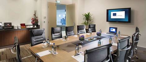 A conference room with a long table, black leather chairs, a TV on the wall, and a reception desk with a plant.
