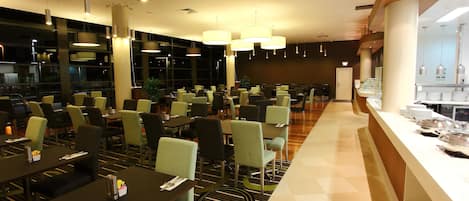 Restaurante moderno com um balcão comprido, várias mesas e cadeiras. Há luminárias pendentes e uma grande janela ao fundo.