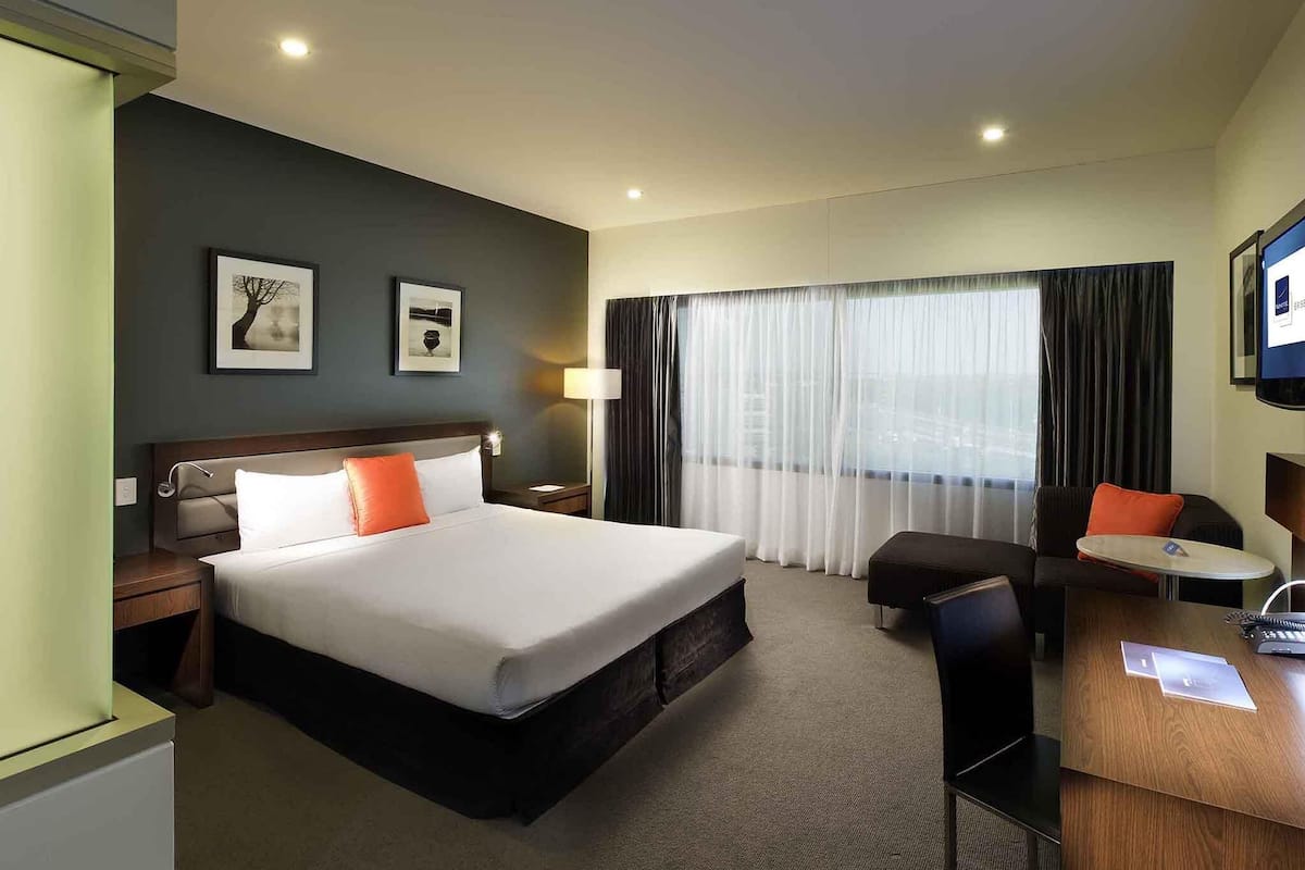 Une chambre d’hôtel avec un grand lit, un bureau, une chaise, une table de chevet et une fenêtre avec des rideaux.