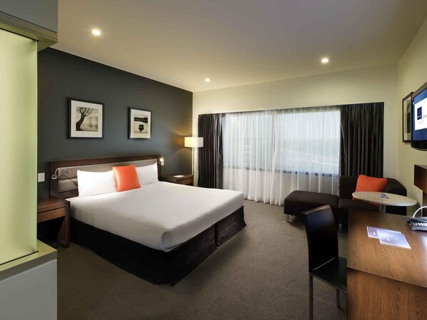 Habitación de hotel con una cama grande, un escritorio, una silla, una mesita de noche y un ventanal con cortinas.