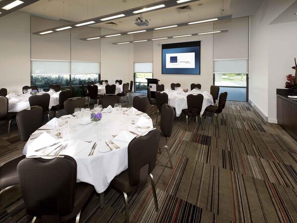 A conference room with round tables, chairs, and a projection screen displaying the Novotel Brisbane logo. - Novotel Brisbane Airport (Brisbane Airport)