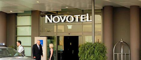 Entrada de un hotel con un cartel que dice 'NOVOTEL', un coche estacionado en frente y una planta en maceta al lado de la entrada.