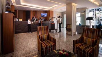 A hotel reception area with a marble floor, striped armchairs, a reception desk, and a television screen.