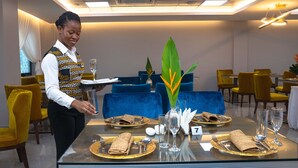 A hotel dining area with a waitstaff setting a table. The room features blue upholstered chairs, yellow chairs, and a glass table. - Oak Plaza Hotels - East Airport (Accra)
