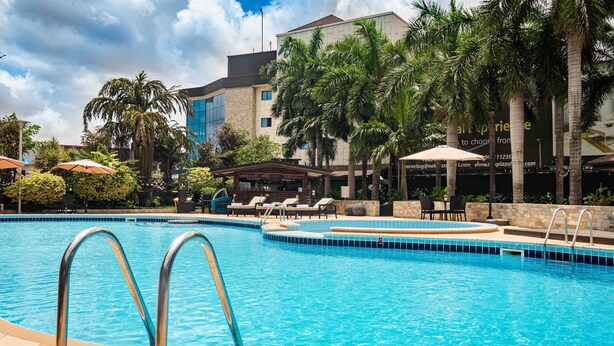 A swimming pool with lounge chairs and umbrellas, surrounded by palm trees and a building with a modern design. - Oak Plaza Hotels - East Airport (Accra)