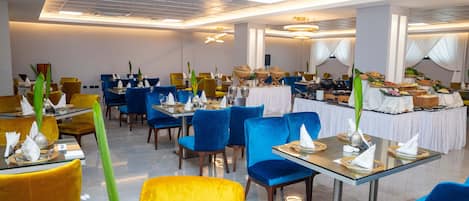 A modern restaurant with blue and yellow chairs, tables set with white napkins, and a buffet area in the background.