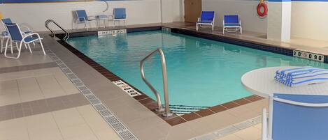 Una piscina interior con paredes de azulejos azules y blancos, señal de zona poco profunda y sillas de descanso alrededor.