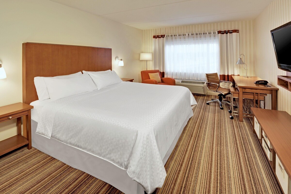 Una habitación de hotel con una cama grande, un escritorio, una silla, un televisor y un ventanal con cortinas.