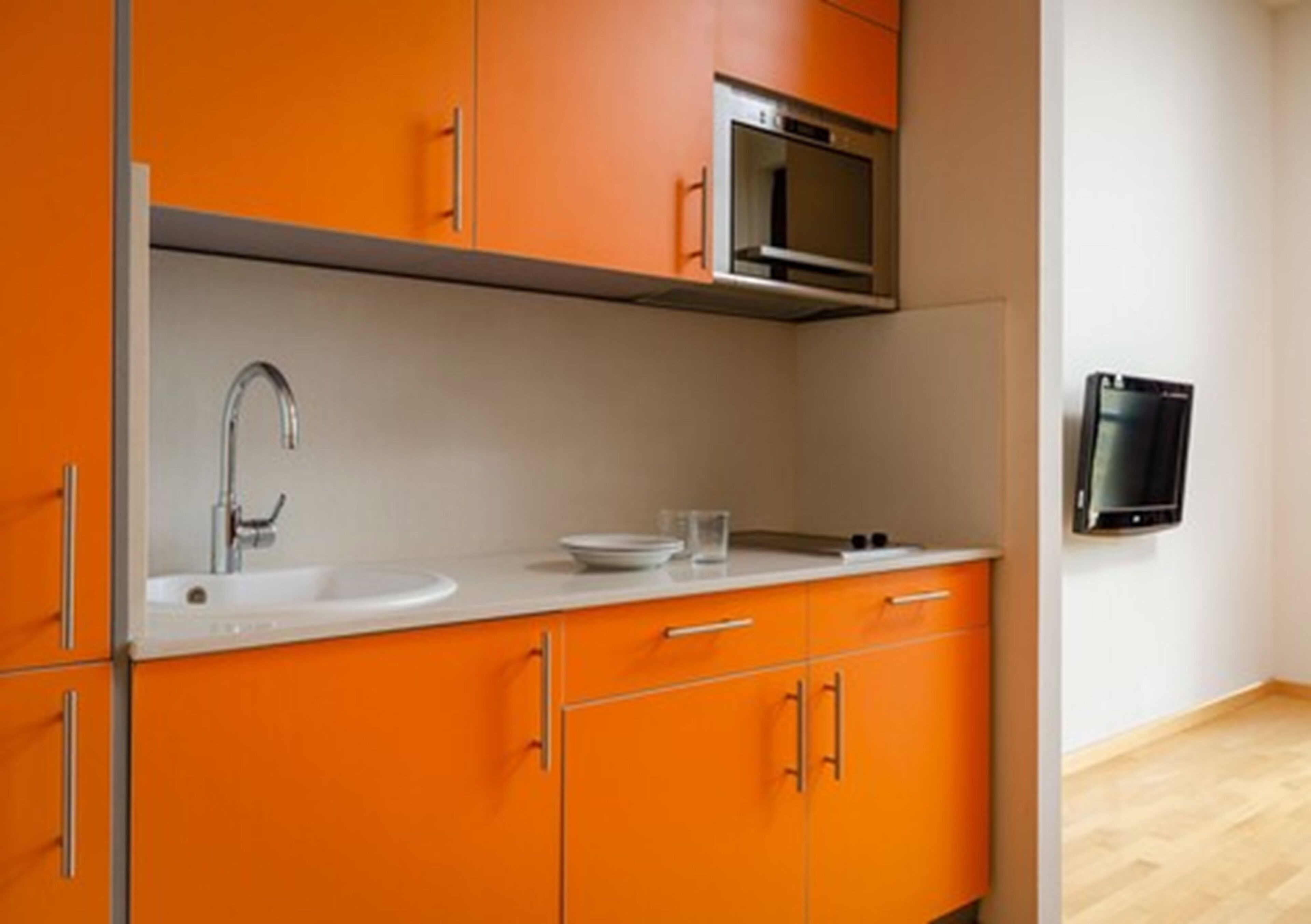 A kitchen with orange cabinets, a stainless steel sink, a microwave, and a flat-screen TV mounted on the wall.