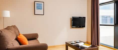 Habitación de hotel con sofá, una mesita con una bandeja de aperitivos, televisor en la pared y ventana con cortinas.