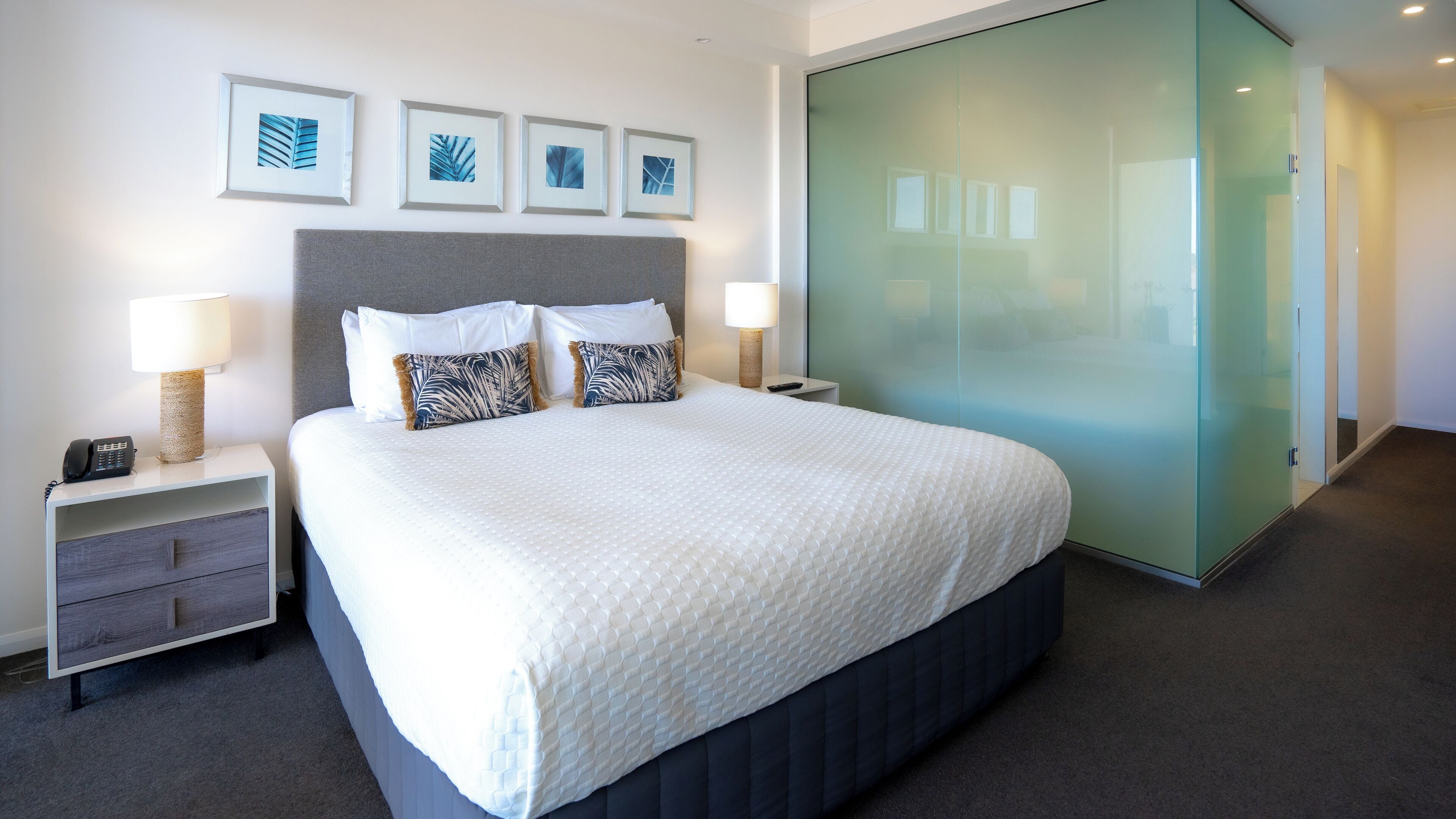 A modern hotel room with a large bed, two bedside tables, a nightstand, a glass partition wall, and framed artwork on the wall.